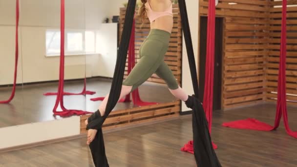 年轻的黑发女孩在一个现代的健身房里 带着木制的隔板和一面镜子 用深色的吊床做着飞舞瑜伽 笑着看着 做着惊人的舞蹈特技表演 穿着裸体的颜色 — 图库视频影像