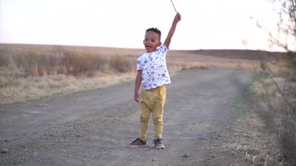 在南非冬季黄昏时分 一个皮肤黝黑的小男孩背着一根干手杖走在一条灰色的砾石路上 — 图库视频影像