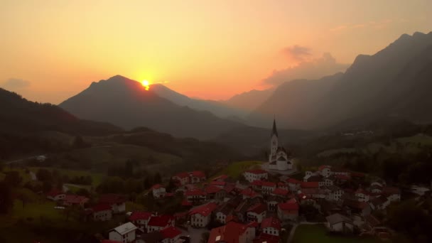 これはスロベニアのコバドリの左から右へ飛ぶ4Kドローンです 背景には教会や山の裏に沈む夕日が見えます — ストック動画