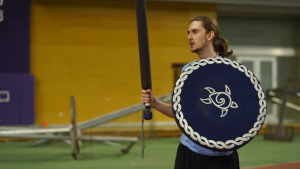 一个留着长发的年轻人带着盾牌和剑在体育馆里指挥Larp泡沫战斗机班 — 图库视频影像
