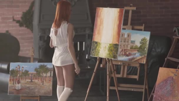 スレンダーな若い赤髪の女性は彼女の作業室で唯一の白い透明シャツに立っているとイーゼルに絵を描く — ストック動画