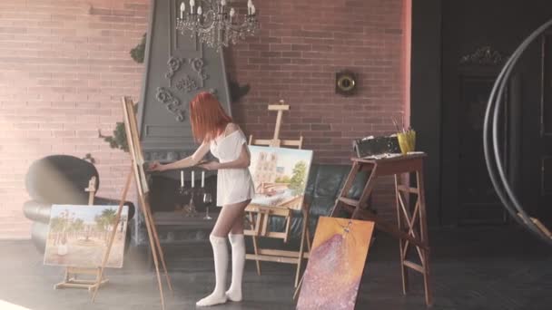 セクシーな若い赤髪の女性は白い透明なシャツと絵画だけで彼女の作業室に立っている 客室はロフトスタイルで作られています — ストック動画