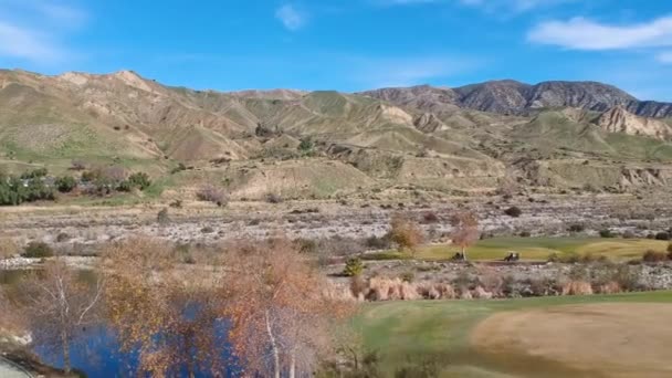 在一个美丽的高尔夫球场上 一个钻机从灌木丛后面升起 看到山峦和3辆高尔夫球车沿着小路疾驰而过 — 图库视频影像