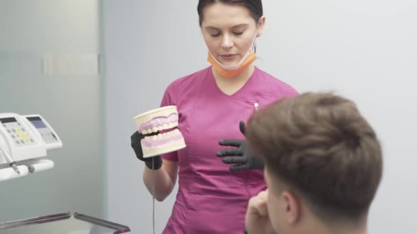 人間の歯の人工モデルを用いた男性患者の手順と治療について説明する手に彼女の制服と手袋の女性歯科医 — ストック動画