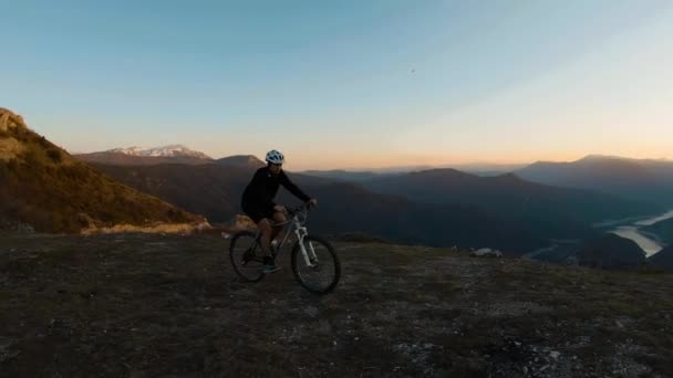 若い男が山の頂上で自転車に乗って夕日を背景に渓谷の湖と — ストック動画