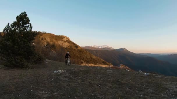 若い男が日没時に山の頂上で自転車に乗っている 背景にある渓谷の湖 — ストック動画
