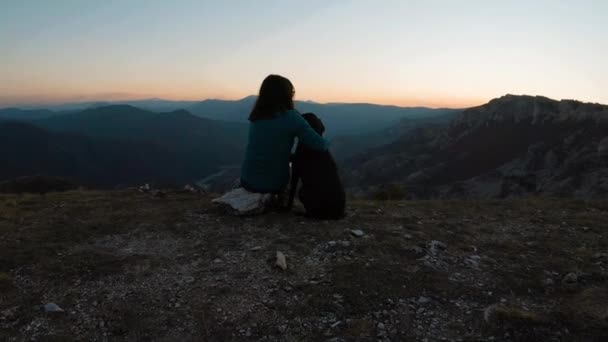 女の子が座っていると山の上に黒いラブラドール犬を抱っこ 背景の夕日の美しい湖の峡谷 後ろからの眺め — ストック動画