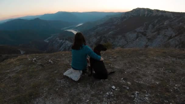 女の子が座っていると山の上に黒いラブラドール犬を抱っこ 背景の夕日の美しい湖の峡谷 後ろからの眺め — ストック動画