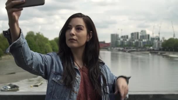 美しいラティーナは ロンドンの景色を望む橋の上に立っている間 彼女の携帯電話で自撮りをしながら ポーズをとって素晴らしい時間を過ごしています — ストック動画