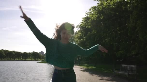 ロンドンの公園で湖の隣で踊るかわいいイタリアの女の子は ジャンプして手を動かします 楽しいです楽しいですフリーながら興奮した女の子 — ストック動画