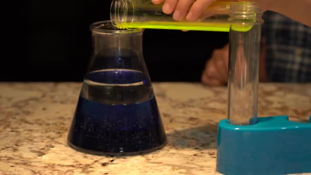 将荧光绿色液体物质从塑料试管倒入装有蓝水和婴儿油的烧杯中的小手 然后他关了灯 你可以看到烧杯的光芒 — 图库视频影像