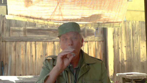 60代の大人の男性が葉巻を吸って日陰の布の下に屋外に座っている姿 — ストック動画