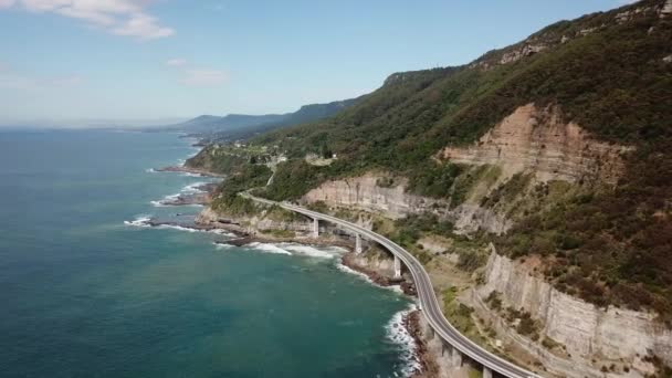 オーストラリアのニューサウスウェールズ州で晴れた日の間に車が横断する際の車の運転として 海の崖の橋の横に沿って高高度ドローンショットトラッキング — ストック動画