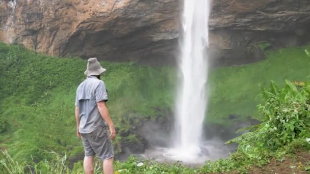 熱帯アフリカの強力な滝の底に立っているブッシュマンの帽子をかぶった髭の生姜男 — ストック動画