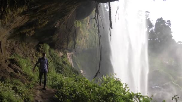 熱帯の滝の後ろの洞窟を歩くアフリカ人の男のスローモーションショット — ストック動画