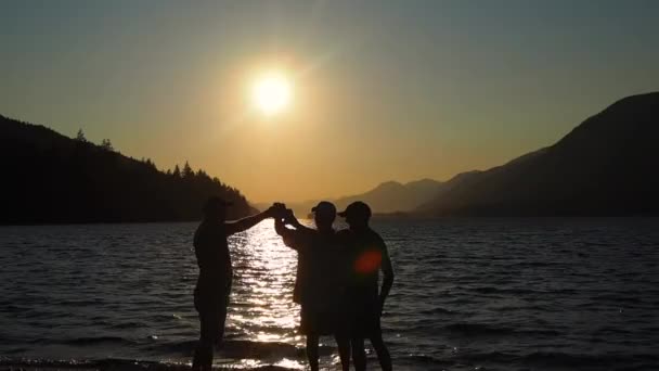 三人の男がシルエットで酒を飲んでいた背景に山のある湖の夕日 — ストック動画