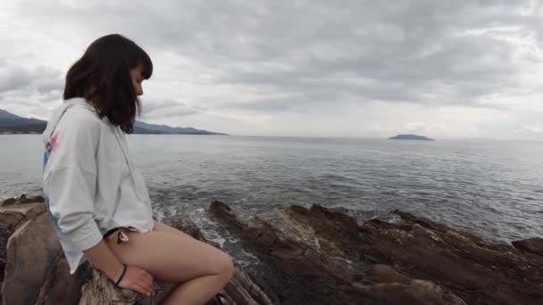 希腊一个阳光明媚的日子里 一位20多岁的黑发女子独自坐在海滨岩石上 — 图库视频影像