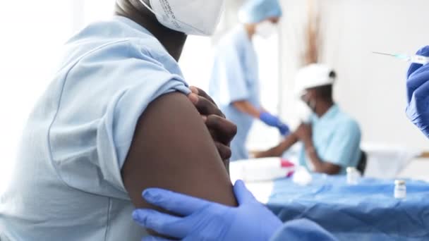 看護師管理者コヴィド 19左腕に注射されたワクチンはクリニックで医療マスクをした黒人男性の綿で腕を拭きます ミディアムショット — ストック動画
