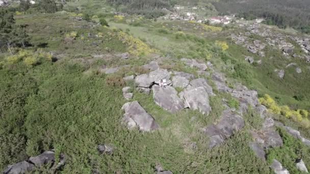 明るい晴れた日に丘陵地帯に囲まれた丘の上に岩の空中回転ショット スペインのガリシアに緑の植生を持つ丘の上の白い椅子 — ストック動画