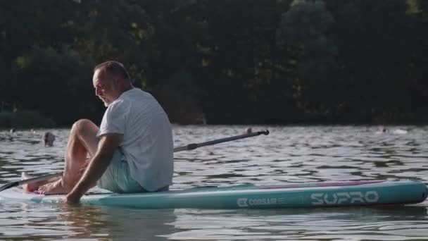 夕日の光の中で湖の真ん中にあるパドルボードに座っている老人は 楽しい瞬間を楽しんでいます — ストック動画