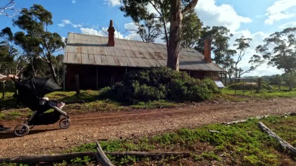 未舗装の道を歩く女性 ビクトリア地方の古い放棄された遺産の錫の屋根の家を通り過ぎるベビーカーを押す — ストック動画