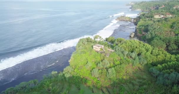 崖の上の民家でインドネシアの緑の海岸線に到達する波の軌道ドローンショット 木々に囲まれた家 — ストック動画