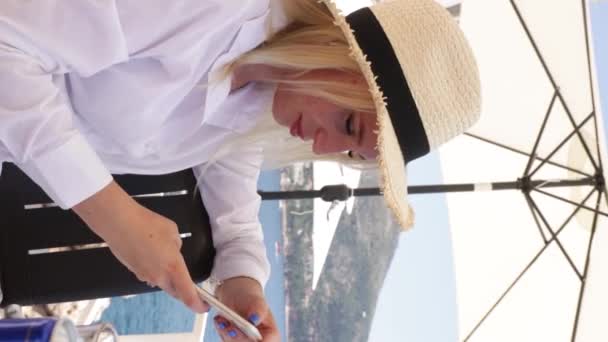 一位年轻漂亮的女士正在海边的一家餐馆里用她的智能手机 穿着白色长袖衬衫 头戴草帽 — 图库视频影像