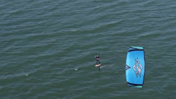 在平静的水面上 一个人滑行水翼风筝并与蓝色帆一起疾驰而过的跟踪航拍 — 图库视频影像