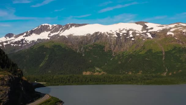 夏には青空に対して部分的に雪をかぶったアラスカの山々の空中風景 湖で山の麓高速道路の道路 サイドドリーショット — ストック動画