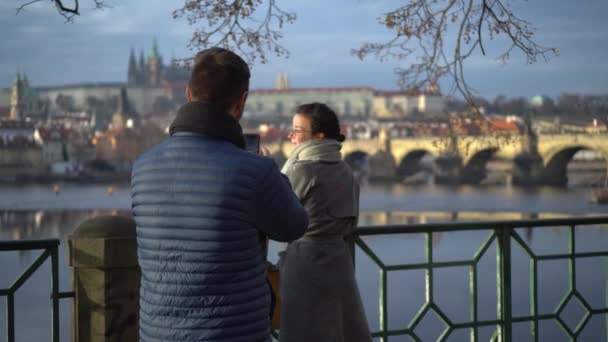 プラハ城とカレル橋の前でガールフレンドの写真を撮る彼氏 携帯電話の写真 — ストック動画