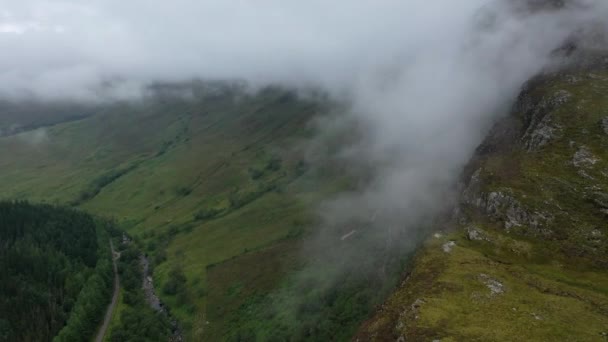 壮大な雲に覆われたピークを越えて木に覆われた谷に来るスコットランドの高地で撮影されたドローン — ストック動画