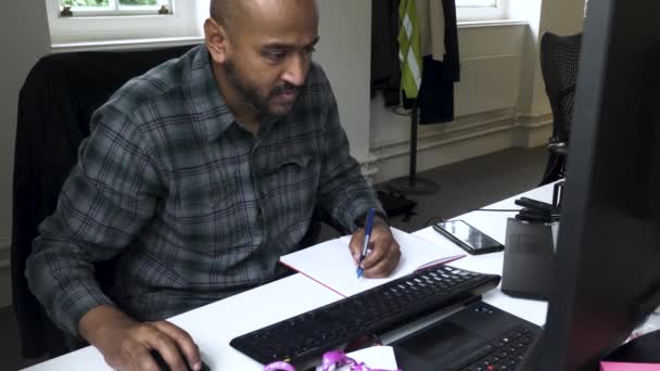 彼のオフィスで働いている茶色の男のショットと彼のコンピュータからいくつかのメモを書き留めた — ストック動画