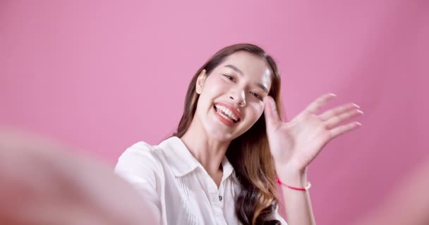 ピンクの背景の前に立ち挨拶や挨拶で手を振ってカメラを迎えてくれる美しいアジア人女性 — ストック動画