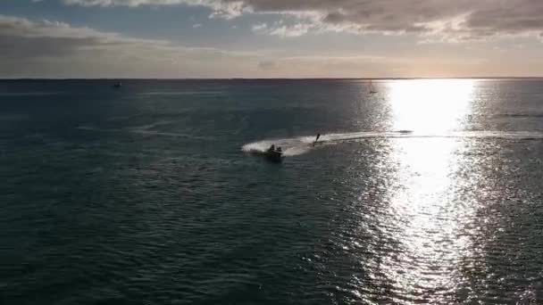 日没時に太陽の水の反射の前の曲線を描写する水上スキーの空中撮影 背景には小さなスポーツヨットがあります — ストック動画