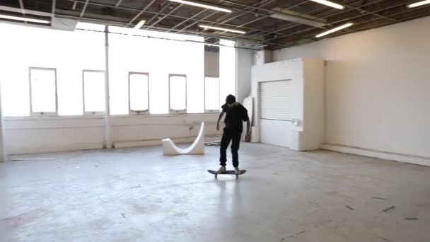 黒い服を着た若い男のスローモーションビューと放棄された空の工場の建物で彼のスケートボード上で運転 — ストック動画