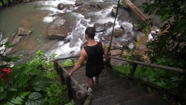 一名身穿黑色衣服的年轻而健康的男子走下台阶和楼梯 走向巴厘岛茂密的绿色丛林中的一条河流 自己探索 环视大自然 — 图库视频影像