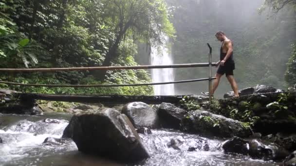 巴厘岛一座横跨河流的竹桥上 一个年轻 白种人正在穿过一座手工建造的竹桥 美丽而巨大的瀑布背景 以慢动作从右到左交叉 — 图库视频影像
