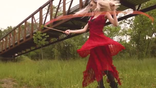 赤いドレスの若いブロンドの女性はリボンの杖を巻きながら牧草地の周りで踊ります シネマティック4Kスローモーション — ストック動画