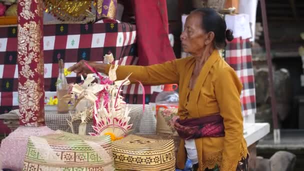 バリのヒンドゥ教の寺院では 高齢の女性は古い野生の花を供え物から取り除く傾向があります 華やかなウィッカーバスケットとカラフルな赤いバナーと装飾 — ストック動画