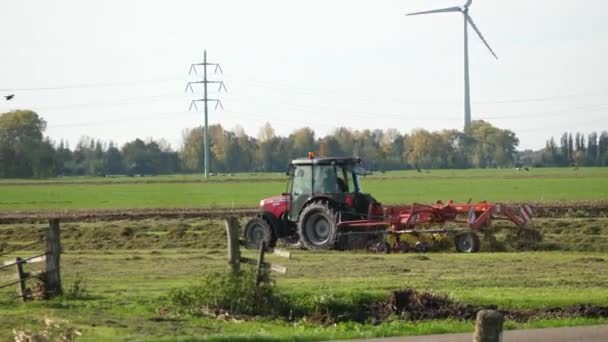 赤いトラクターは オランダの晴れた秋の日に畑で作物を切っています 遠くの大きな風力タービンと電力線の背景 — ストック動画