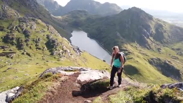 一名年轻的女徒步旅行者正走在挪威罗浮敦的豪克兰海滩附近的曼宁山上 在温暖的夏日 她在阳光下快乐地微笑着 背后是美丽的湖泊和高山 — 图库视频影像