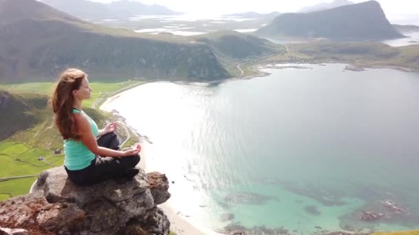 一名年轻的白人女徒步旅行者正在曼宁山顶上悬崖边的岩石上坐着沉思 在挪威罗浮敦一个温暖而阳光明媚的夏日里俯瞰着霍克兰海滩 — 图库视频影像