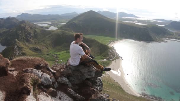 一个纹身的年轻人正一个人坐在那里 看着下面的豪克兰海滩的美丽景色 调整他的姿势 使他更舒服地坐在挪威罗浮敦的山顶上 — 图库视频影像