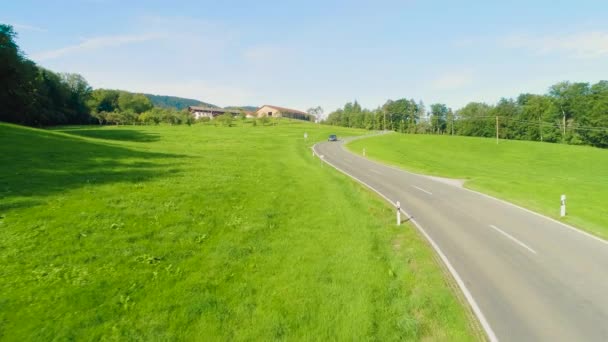 丘の上の家と晴れた日に明るい緑の芝生と丘陵地帯の道路上の車の運転の低い航空フロントビュー — ストック動画