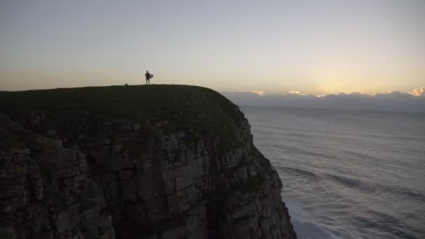 海によって日の出に崖の上にサーフボードを持つ男 — ストック動画
