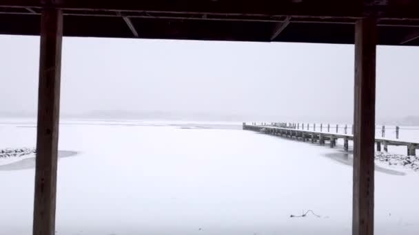 雪の中で木製のカバナの下を飛んで凍った白い川と雪に覆われた桟橋に出てくる — ストック動画