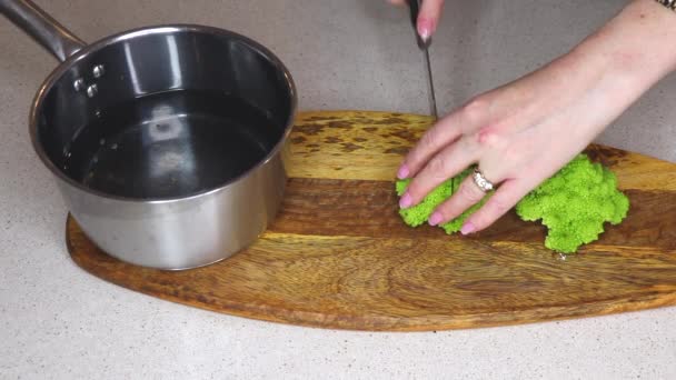 新鮮な赤ちゃんのロマネスコブロッコリー カリフラワーを木製のまな板に鋭利なナイフで切る女性料理 水の鍋にみじん切り作品を追加します — ストック動画