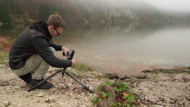 写真家は 湖の縁の横に写真を撮るために地面の近くに位置三脚に彼のカメラを設定します — ストック動画