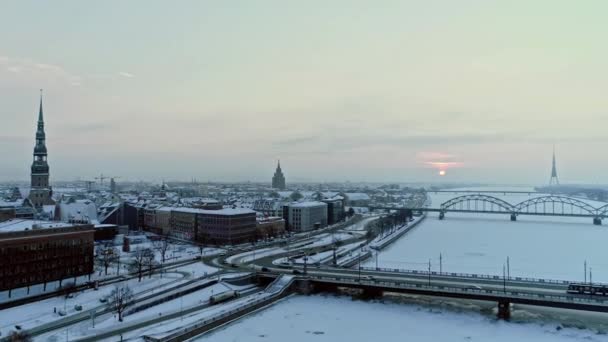 午前中の雪の多い冬の日のリガ市内の空中逆撮影 フローズン ダウガヴァ川 運転車 日の出中の大聖堂 ラトビアのリガ首都 — ストック動画
