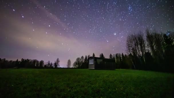 空を飛ぶ星や流星と森の上の空と芝生のフィールド上の照明コテージで美しい夜の時間の経過 — ストック動画